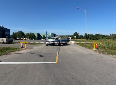 Wide view of a road barrier at a construction site with a white utility truck and industrial buildings in the background under a clear blue sky