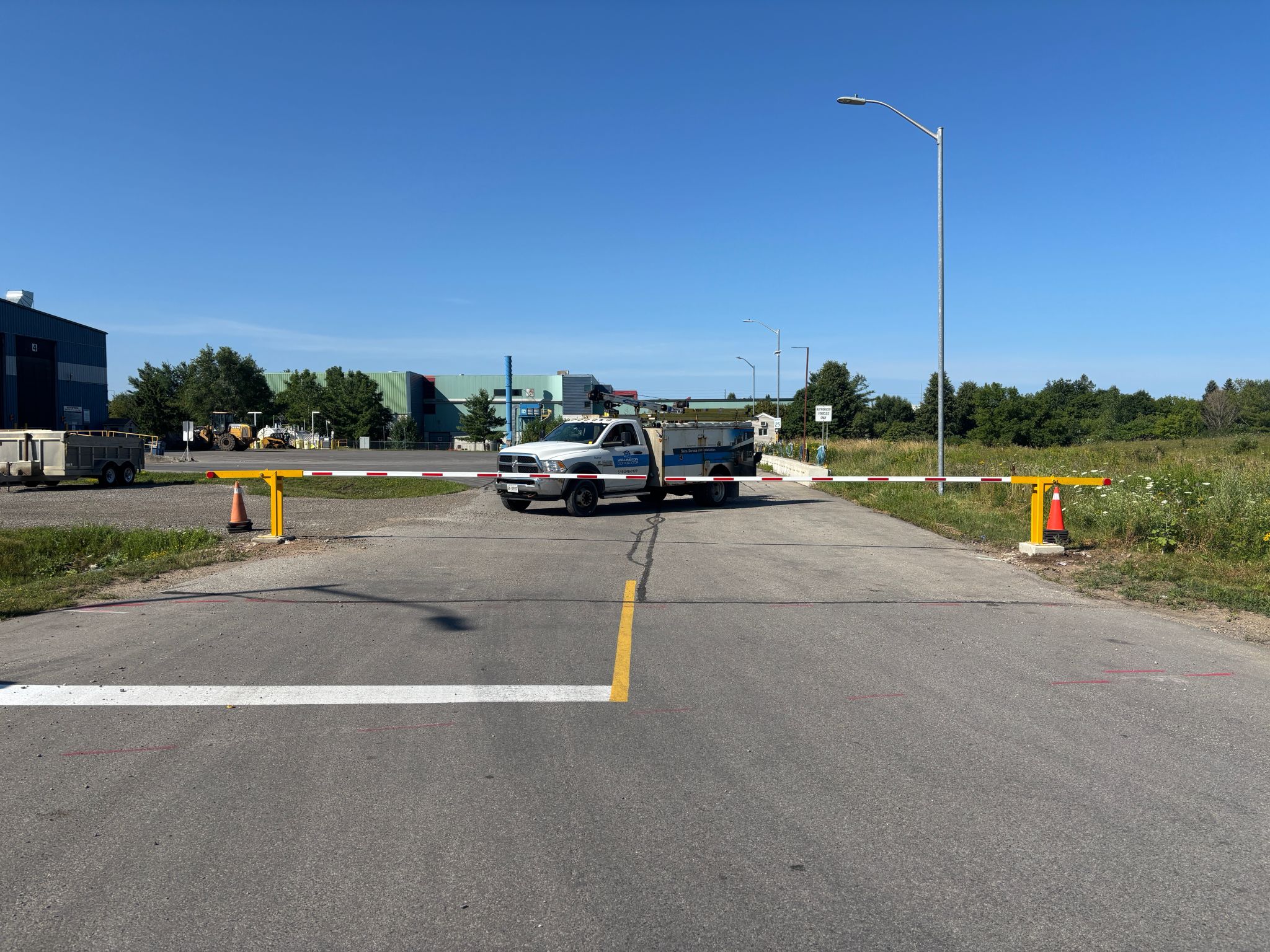 Wide view of a road barrier at a construction site with a white utility truck and industrial buildings in the background under a clear blue sky