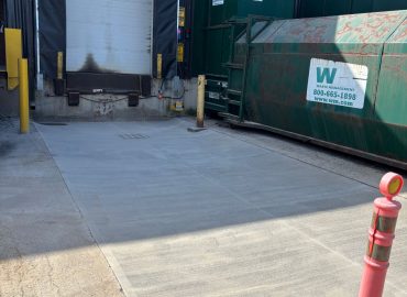 Loading dock area with a closed roll-up door and large green Waste Management dumpsters in front; an orange safety cone is in the foreground.