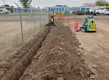 Long trench along a chain-link fence at a construction site, with a small excavator, red safety tape, and portable toilet in the background.