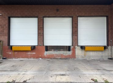 Three white roll-up garage doors in a red brick warehouse, with yellow dock guards along the loading area.