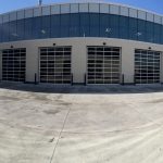 Modern industrial building with a curved glass front and five large garage doors; construction equipment visible on the right in a sunny lot.