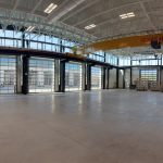Industrial workshop with an overhead yellow crane spanning the bay and multiple large glass roll-up doors along the back wall
