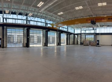 Industrial workshop with an overhead yellow crane spanning the bay and multiple large glass roll-up doors along the back wall