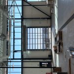 Industrial interior under construction with exposed steel beams, ducts, and a large glass garage door opening to the outdoors at the far end of the space