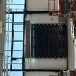 Industrial warehouse interior with a large dark sectional garage door, exposed white beams, and scattered cardboard boxes and pallets on the concrete floor.