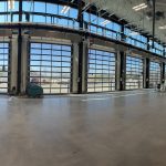 Spacious warehouse with tall glass garage doors along the wall, scattered equipment and boxes inside.