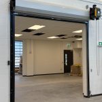 Open industrial loading bay with a large overhead door raised to reveal a warehouse interior and pallets nearby