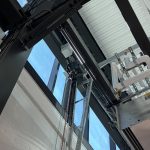 Industrial mechanical ceiling with black steel beams, exposed cables and white pipes near a blue windowed wall.