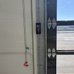 Interior view of a garage door opening with a LiftMaster control panel mounted on a metal track, chain hanging beside a white cinder block wall and red/green wires visible