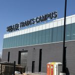 Modern building with a 'Skilled Trades Campus' sign on the roof, under construction with pallets and portable toilets in the foreground.