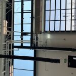 Industrial interior with exposed white ductwork, piping, and a large glass garage door letting in daylight outside.