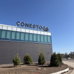 Building with a large 'CONESTOGA' sign atop, against a clear blue sky; foreground shows a dirt lot, orange safety cones, and a new curb work area.