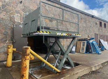 Green hydraulic scissor lift raised near a stone facade building, with yellow safety bollards in the foreground and pallets nearby